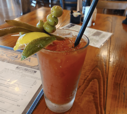 A colorful cocktail garnished with olives, lemon, and pickles, served in a glass on a wooden table.