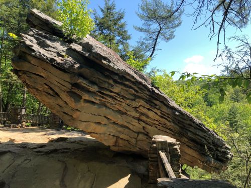 A large, tilted rock formation surrounded by trees and greenery, with a clear blue sky in the background.