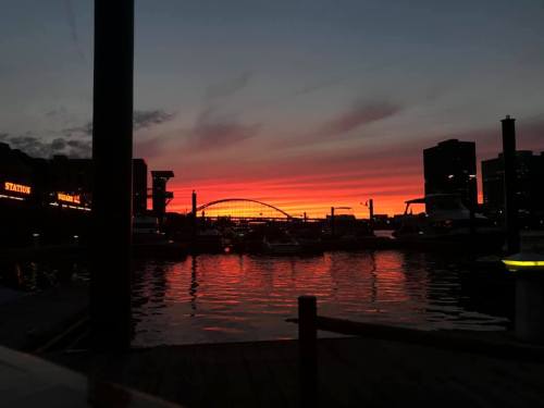 A vibrant sunset over a harbor, with silhouettes of buildings and boats reflecting on the water.