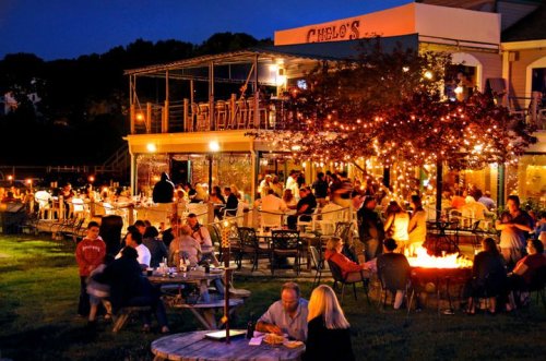 A lively outdoor restaurant at night, filled with people dining and enjoying the ambiance of string lights and fire pits.