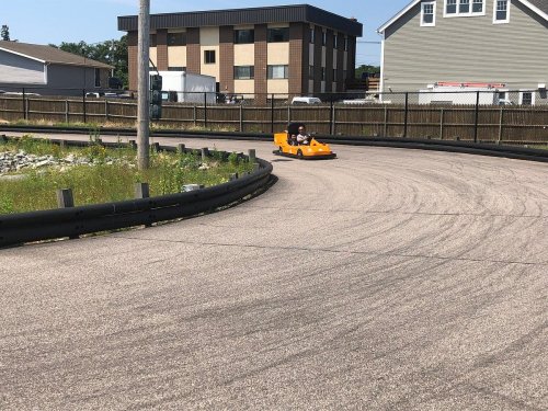A person drives an orange go-kart on a curved track, with buildings and greenery in the background.