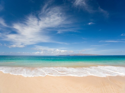 A serene beach scene with gentle waves, soft sand, and a clear blue sky with wispy clouds.