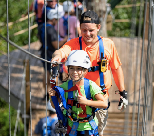 A child in a harness smiles as an adult guides her across a suspension bridge in a forested area.
