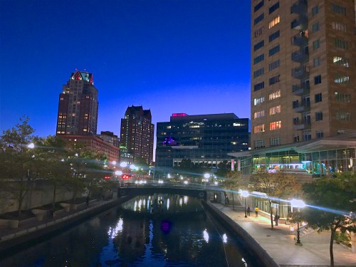 City skyline at dusk, featuring buildings reflecting in a calm river, with lights illuminating the waterfront.