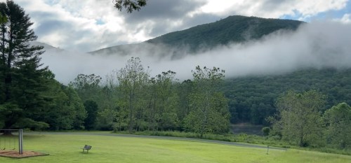 A serene landscape featuring mountains shrouded in mist, surrounded by lush greenery and a grassy area in the foreground.
