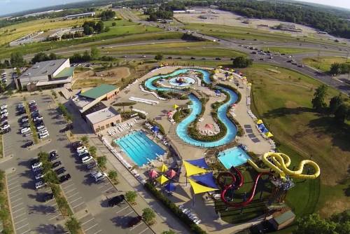 Aerial view of a water park featuring pools, slides, and a lazy river, surrounded by green fields and parking areas.