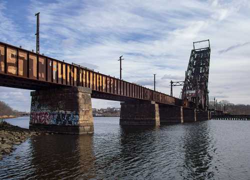 A rusted drawbridge over a river, with graffiti on the stone supports and a cloudy sky in the background.