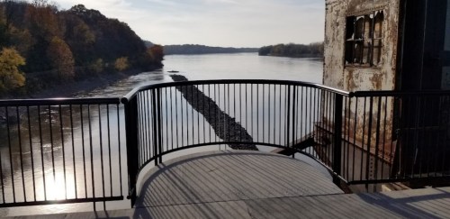View of a calm river with a rocky shoreline, framed by a railing and trees in the background under a clear sky.