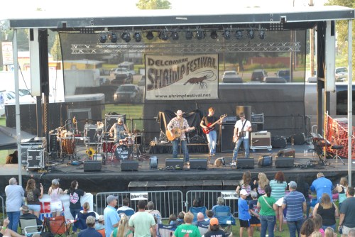 A band performs on stage at the Delcambe Shrimp Festival, with an audience enjoying the show in front.
