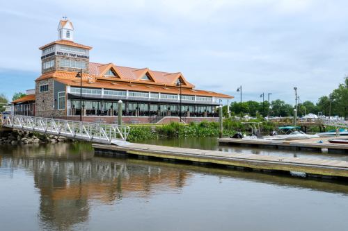 A marina building with a stone facade and orange roof, overlooking a calm waterway with boats and a wooden dock.