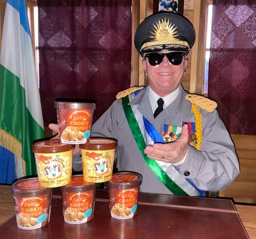 A smiling man in a military uniform and sunglasses poses with five tubs of edible cookie dough.