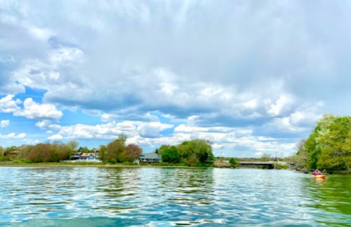 A serene lake scene with fluffy clouds, green trees, and a distant building along the shore.