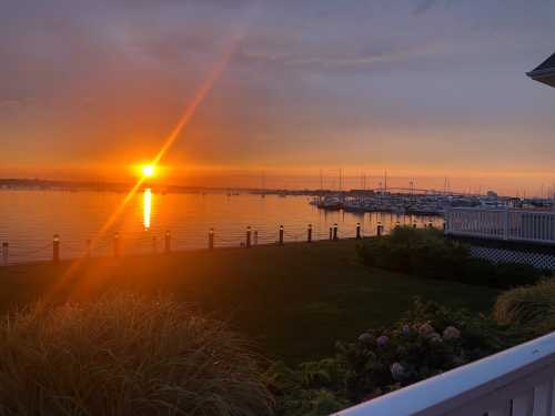 Sunset over a calm harbor, with boats docked and vibrant colors reflecting on the water. Lush greenery in the foreground.