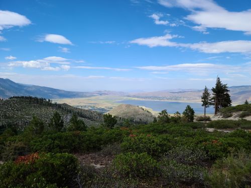 A scenic view of a valley with a lake, surrounded by mountains and trees under a blue sky with scattered clouds.
