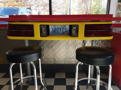 A retro diner bar with a yellow car rear as the counter, featuring a Rhode Island license plate and two black stools.