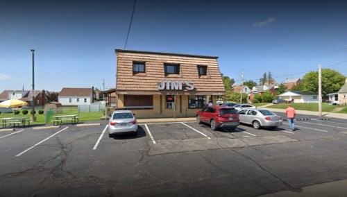 A restaurant named "Jim's" with a brown shingled roof, surrounded by parked cars and a few outdoor tables.