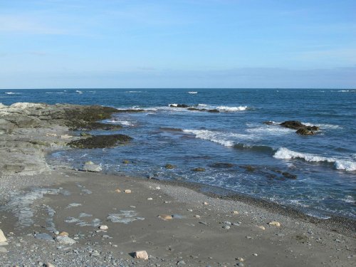 A rocky shoreline with gentle waves lapping at the sand under a clear blue sky.