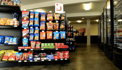 A convenience store aisle filled with various snack bags and drinks, with a checkout area visible in the background.