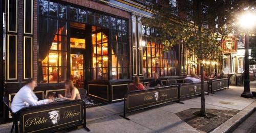 Outdoor dining area of a restaurant with people seated at tables, surrounded by large windows and warm lighting.