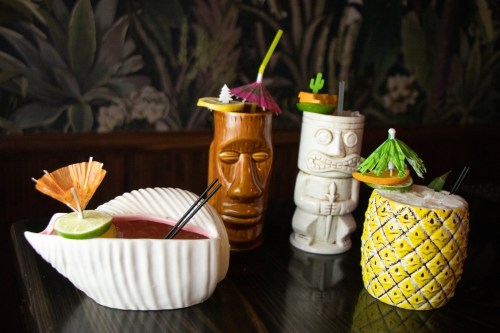 Four colorful tropical cocktails in decorative mugs, featuring fruits and umbrellas, set against a lush background.