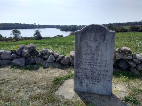 A stone monument near a pond, surrounded by grass and rocks, with a scenic view of the water and trees in the background.