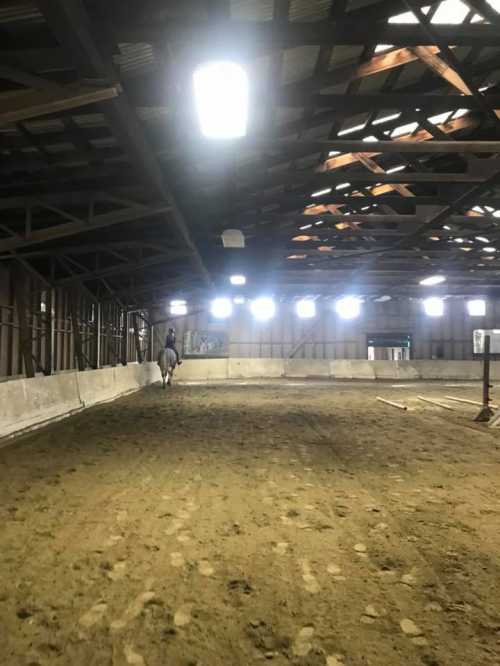 A spacious indoor riding arena with a horse and rider in the distance, illuminated by overhead lights.