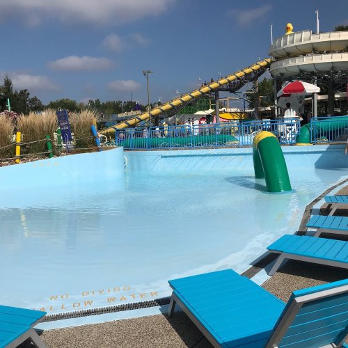 A water park scene featuring a blue pool, lounge chairs, and a water slide in the background under a partly cloudy sky.