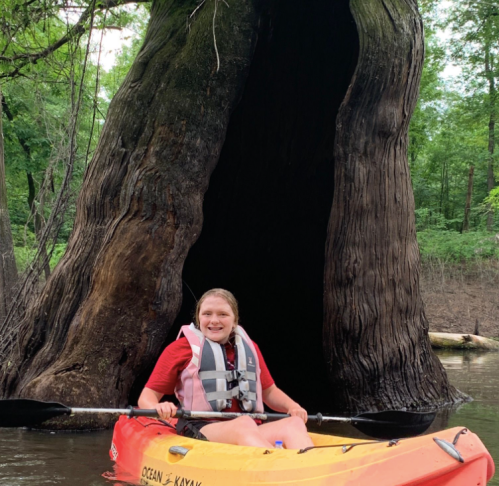 A person in a kayak smiles near a large tree with a hollow trunk, surrounded by lush greenery and calm water.