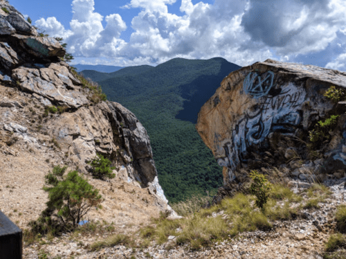 A rocky cliff overlooks a lush green valley under a partly cloudy sky, with graffiti on the rocks.