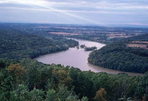 A scenic view of a winding river surrounded by lush greenery and rolling hills under a cloudy sky.