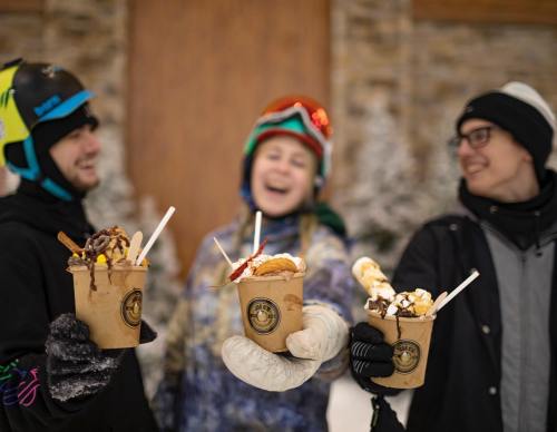 Three people in winter gear hold cups of dessert, smiling and enjoying treats in a snowy outdoor setting.