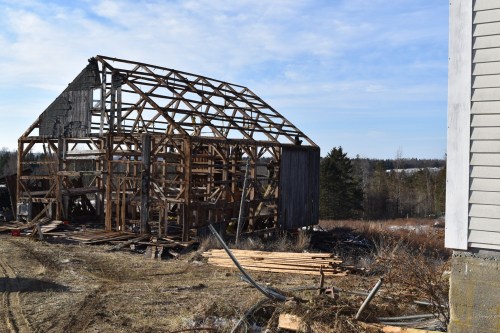 A partially collapsed wooden barn stands in a field, surrounded by debris and trees under a clear blue sky.