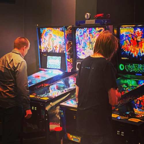 Two people playing pinball machines in a dimly lit arcade, focused on their games.