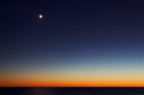 A crescent moon shines in a twilight sky above a calm ocean, with a gradient of deep blue to warm orange hues.