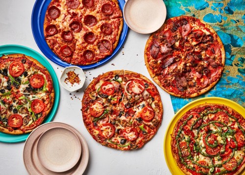 A colorful spread of five pizzas with various toppings, surrounded by empty plates and a bowl of red pepper flakes.
