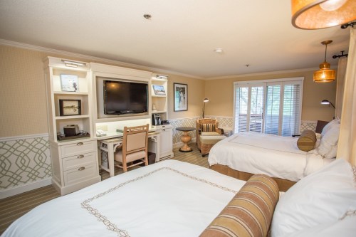 A cozy hotel room featuring two beds, a desk, a TV, and large windows with natural light.