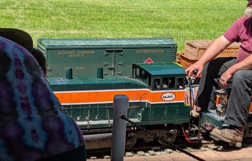 A model train with a green and orange engine, parked next to a green cargo car on a sunny day.
