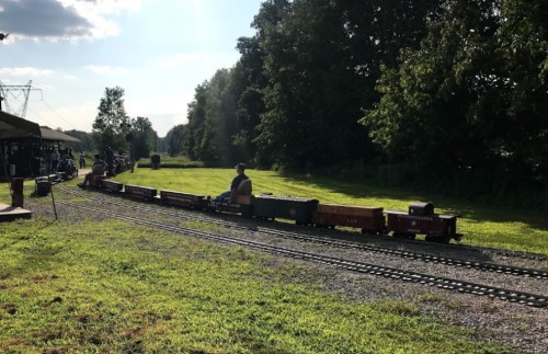 A miniature train on tracks in a green field, with passengers enjoying a ride under a sunny sky.