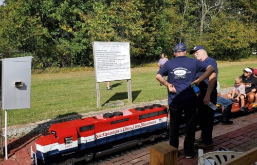 Two men converse near a model train on a track, with a sign and trees in the background. Children sit nearby.