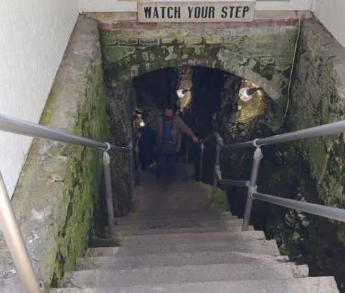 A dimly lit staircase leads down into a moss-covered stone tunnel, with a "Watch Your Step" sign above.