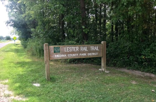 Sign for Lester Rail Trail in Medina County Park District, surrounded by trees and grass.