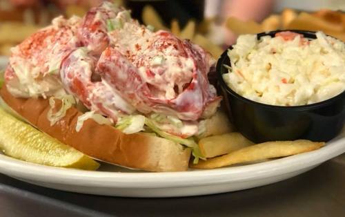 A lobster roll topped with creamy dressing, served with coleslaw and fries on a white plate.