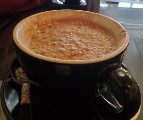 A close-up of a frothy cup of coffee on a black saucer, with a spoon beside it.