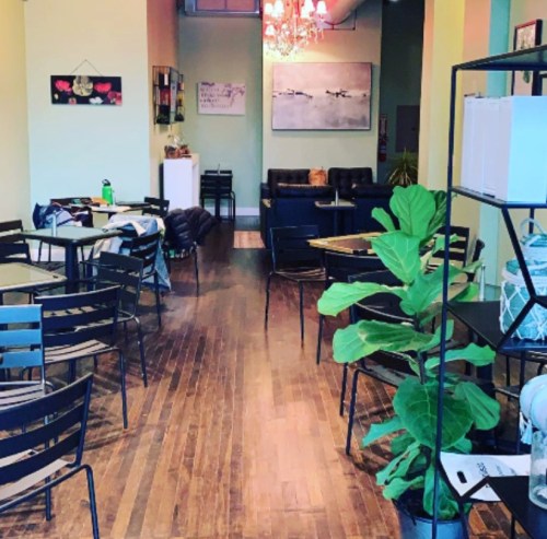 A cozy café interior with wooden floors, empty tables, and a plant, featuring a lounge area in the background.