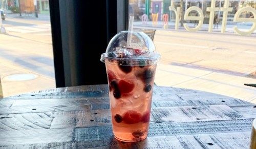 A clear cup filled with a fruity drink, featuring berries and ice, sits on a wooden table by a window.