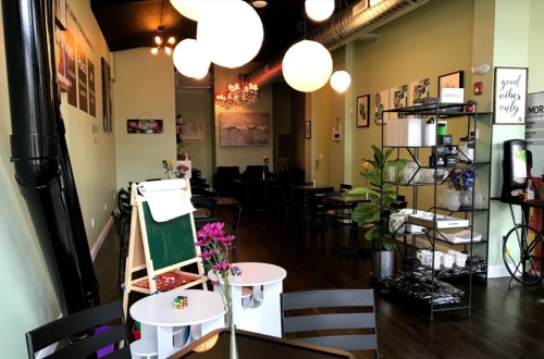 Cozy café interior with round pendant lights, tables, and a display shelf, featuring plants and artwork on the walls.