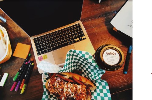 A laptop on a table next to a sandwich in a green checkered basket and a jar of sauce, surrounded by colorful markers.