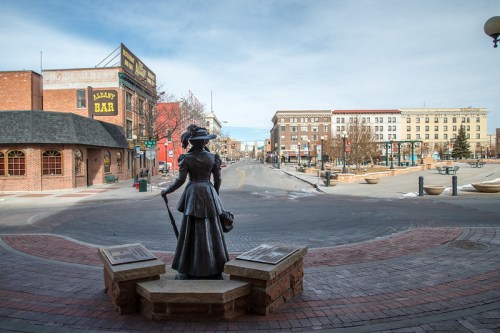A bronze statue of a woman in period dress stands on a brick plaza, overlooking a quiet street lined with buildings.