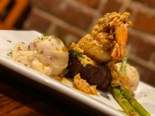A plated dish featuring a steak topped with shrimp, served with mashed potatoes and asparagus against a brick background.