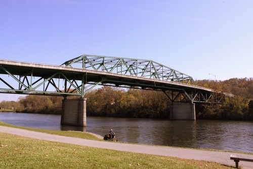 A green metal bridge spans a river, with trees in the background and a path along the water's edge.